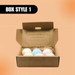 Open cardboard box displaying a "Lush Bath Bomb Gift Set" with three bath bombs nestled in straw. The text reads "Box Style 1" on an orange background.